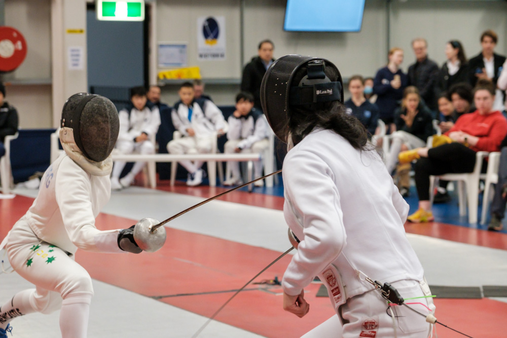 Two fencers on the piste with weapons pointed at each other and a crowd behind them watching